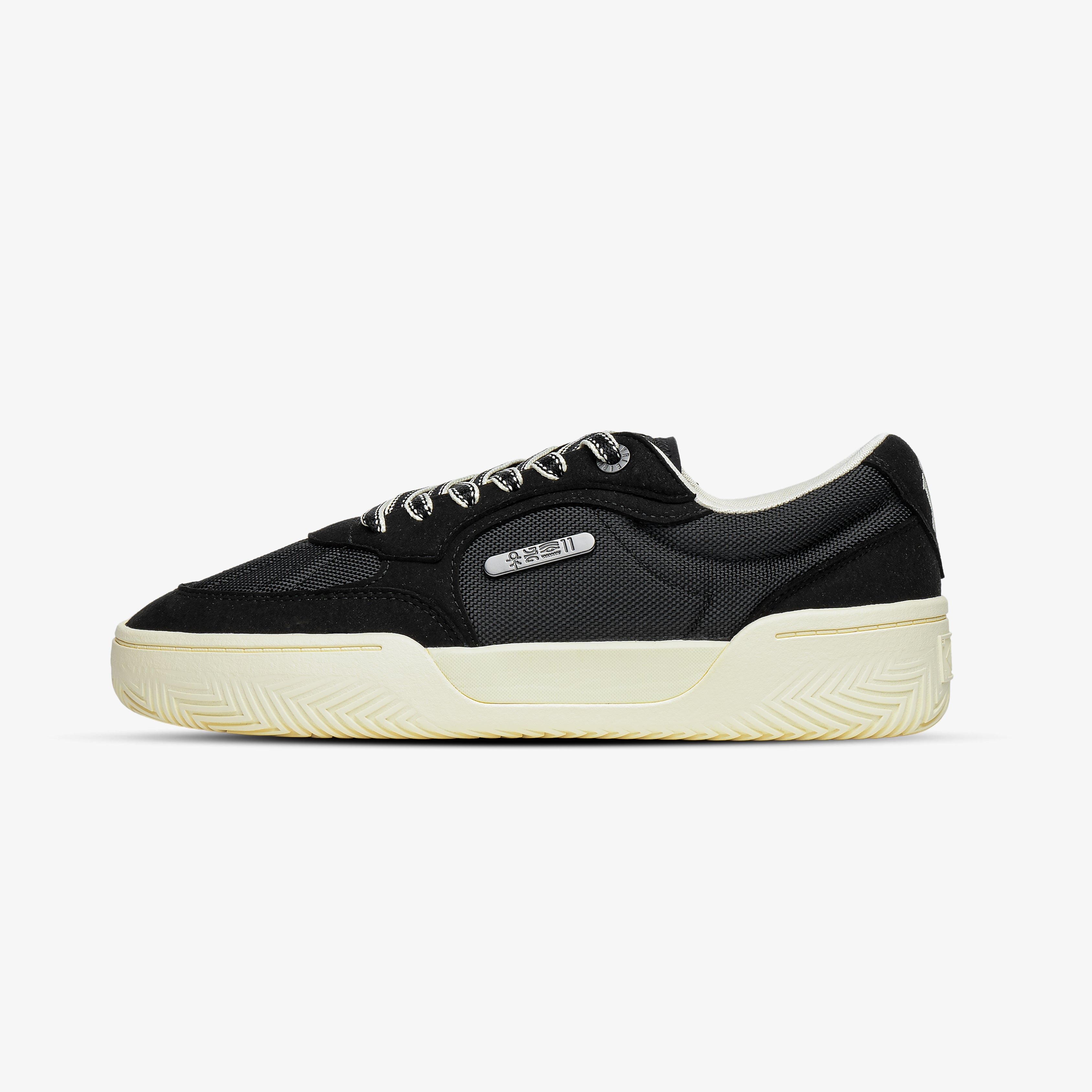 Side view of a black low-top canvas sneaker with cream-colored rubber sole and metallic eyelets, designed in a retro tennis shoe style for men, on a white background.
